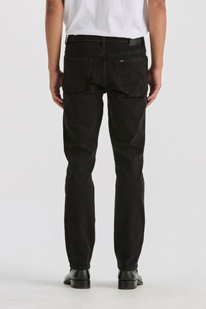 A person stands facing away from the camera against a light background, wearing a white T-shirt, black Lee Originals L100 Slim Jean, and black shoes for a timeless look.