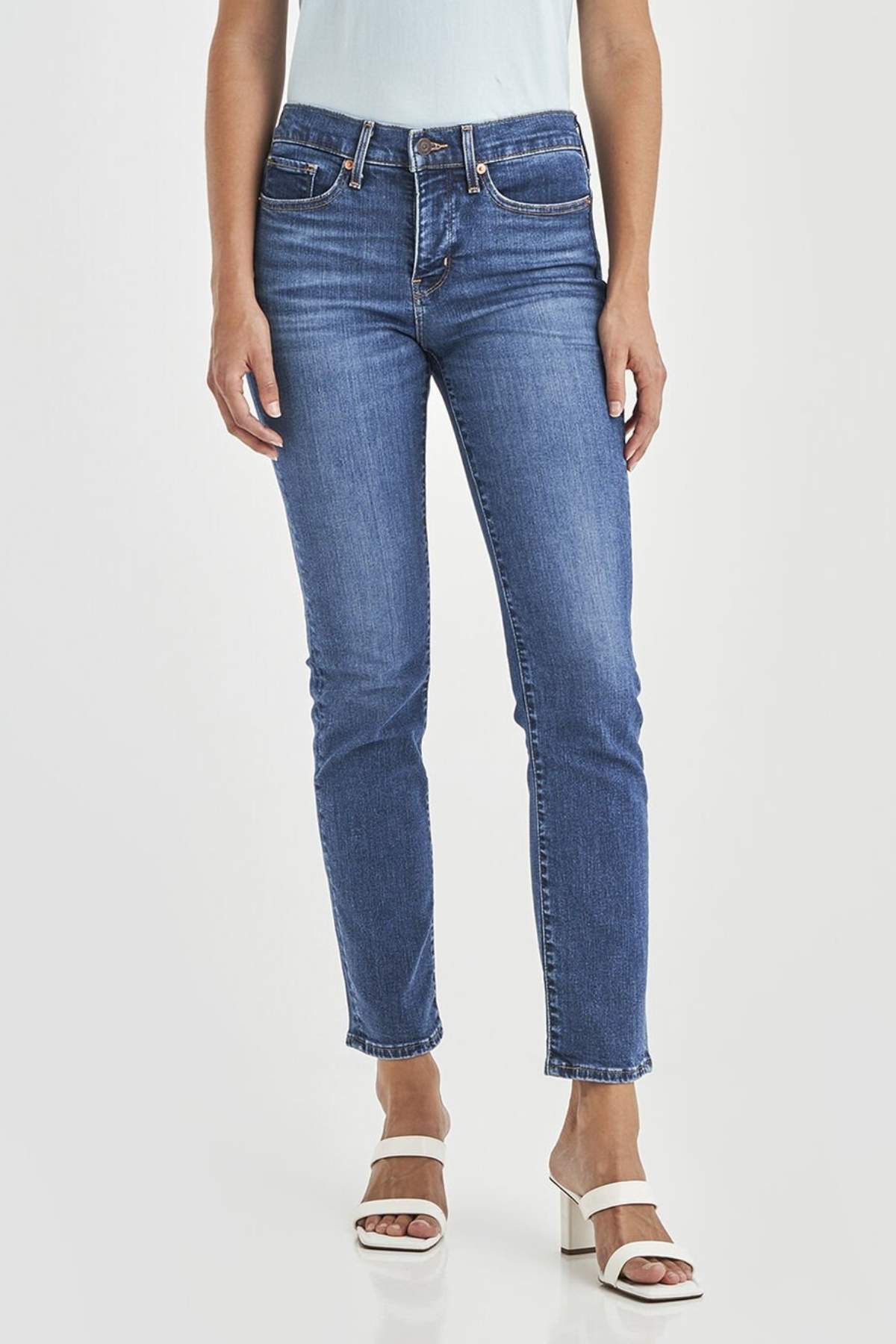 Wearing Levis 312 Shaping Slim Jean in blue stretch fabric and white heeled sandals, a person stands against a plain light background with arms relaxed by their sides.