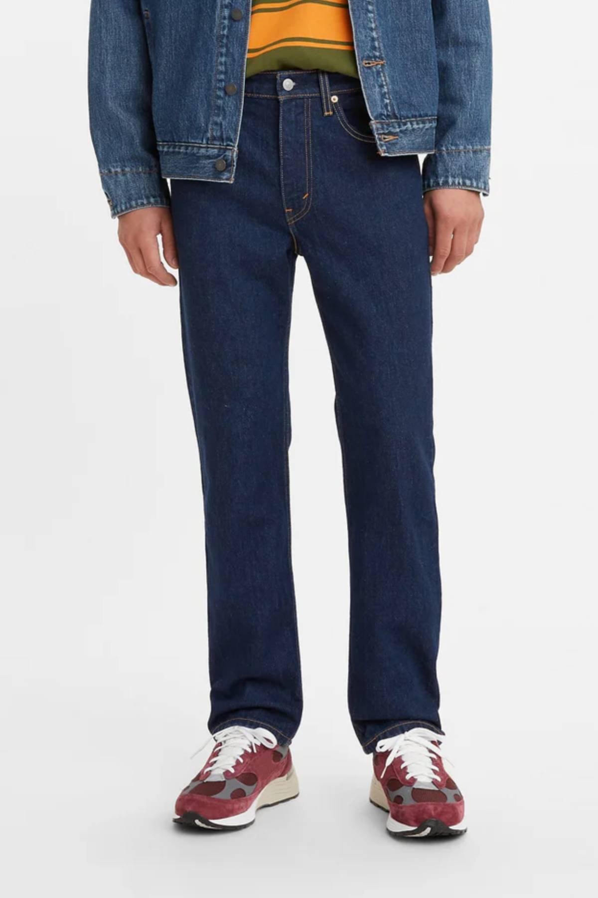A person stands against a white background wearing a blue denim jacket, yellow and green striped shirt, maroon and white sneakers, and Levis 516 Straight Jean in a straight-leg fit.