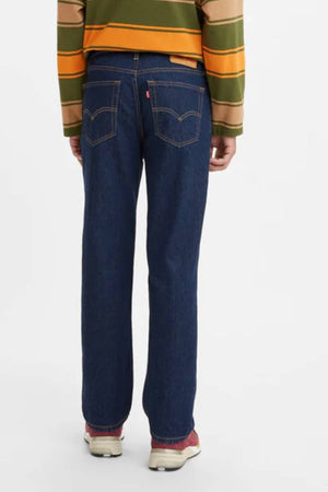 A person stands with their back to the camera, wearing a green, brown, and yellow striped sweater, Levis 516 Straight Jean with a straight-leg fit and visible stitching, and light sneakers featuring red accents on the heels.