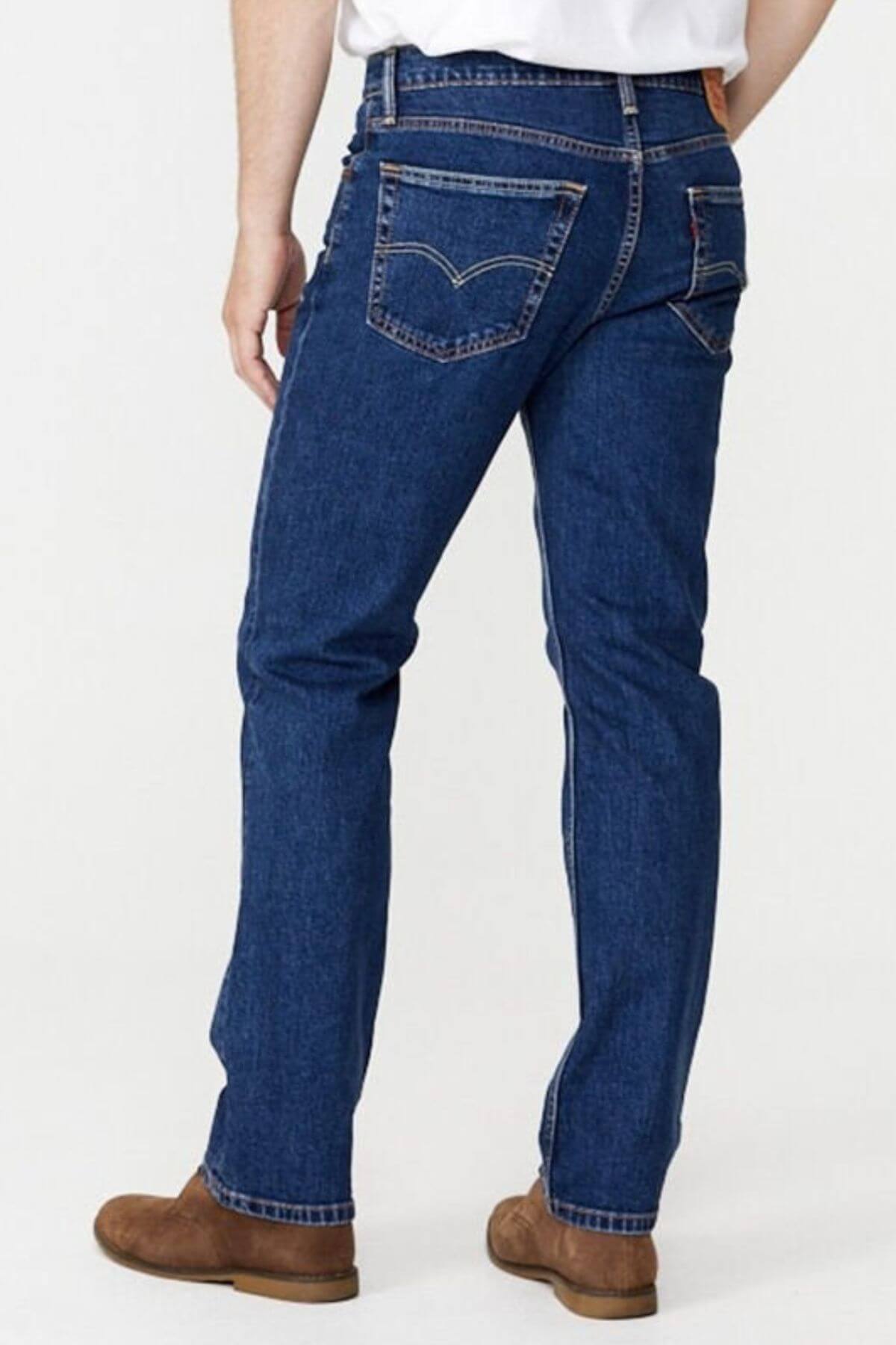 A person standing and facing away, showcasing the back of the 100% cotton Levis 516 Straight Jeans in blue with visible stitching and pockets. The jeans offer a comfortable fit, paired with a white tucked-in shirt and brown shoes. The background is plain and light-colored.