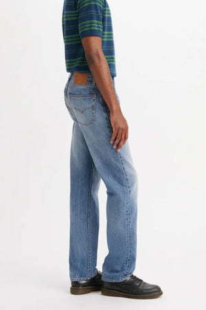 A person wearing a green and blue striped shirt, Levis 555 Relaxed Straight Jean, and black shoes stands sideways against a plain white background.