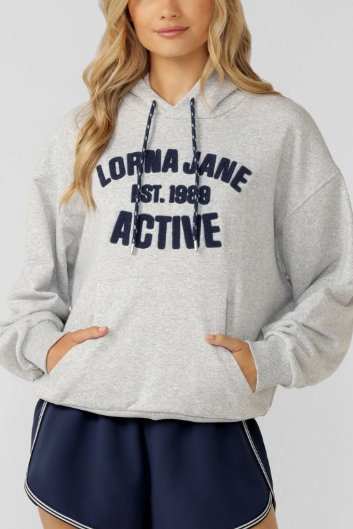 A woman wears a light gray Lorna Jane Active Club Hoodie with "LORNA JANE EST. 1989 ACTIVE" on the front, paired with navy blue athletic shorts featuring white trim.