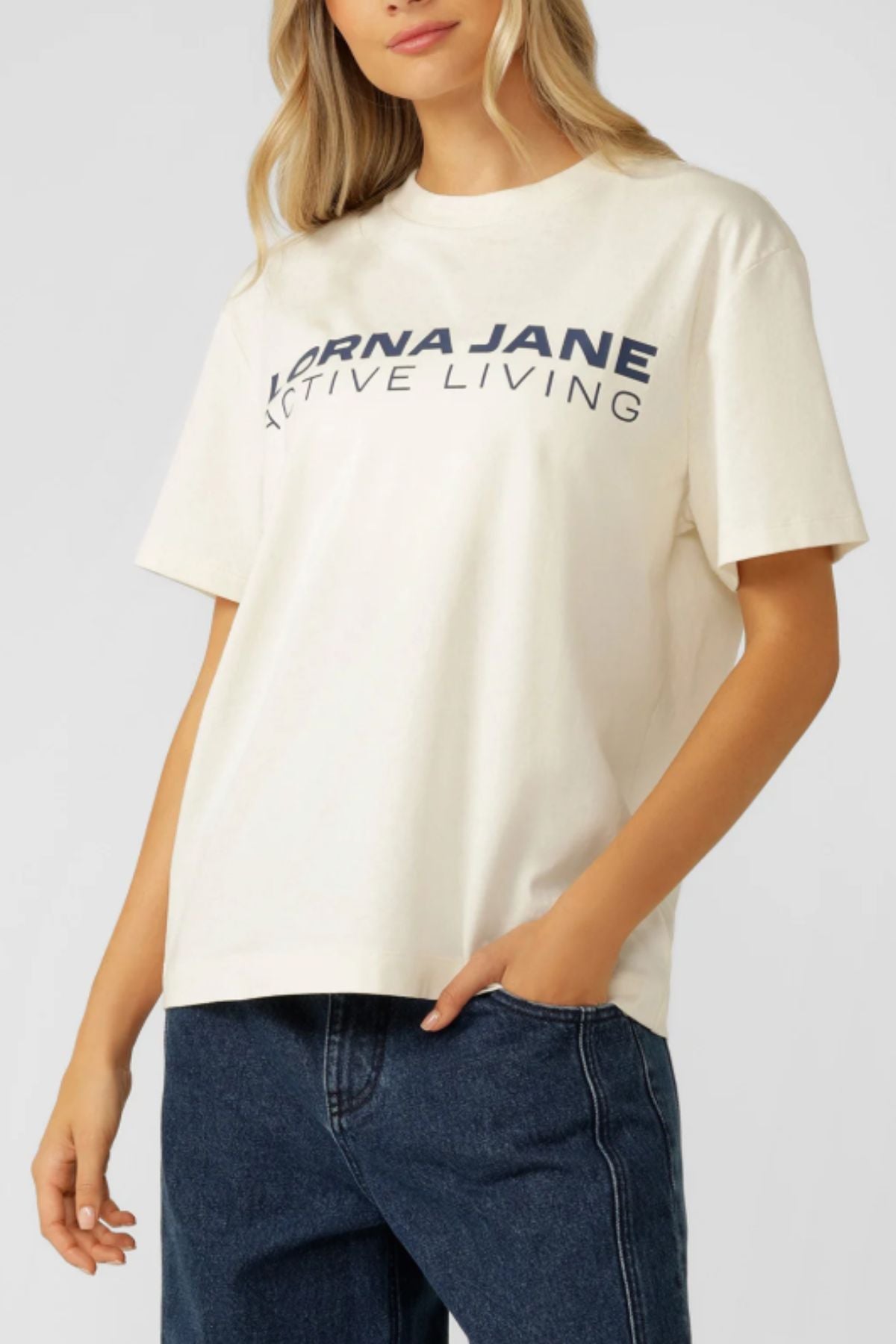 A woman wears the Lorna Jane Active Living T-Shirtโan oversized, premium cotton jersey with bold letteringโpaired with blue jeans, standing with one hand relaxed by her side.