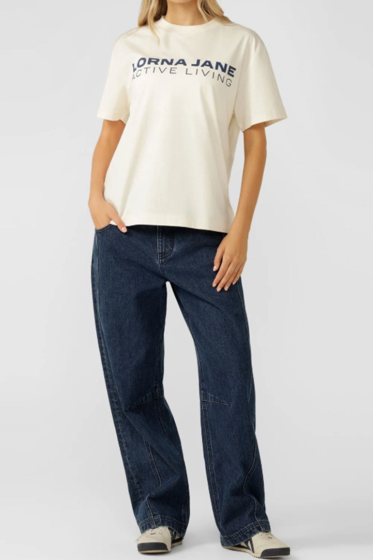 A person stands with one hand in their pocket against a plain background, wearing an oversized cream Lorna Jane Active Living T-Shirt, loose dark blue jeans, and black-and-white sneakersโan essential for active living.