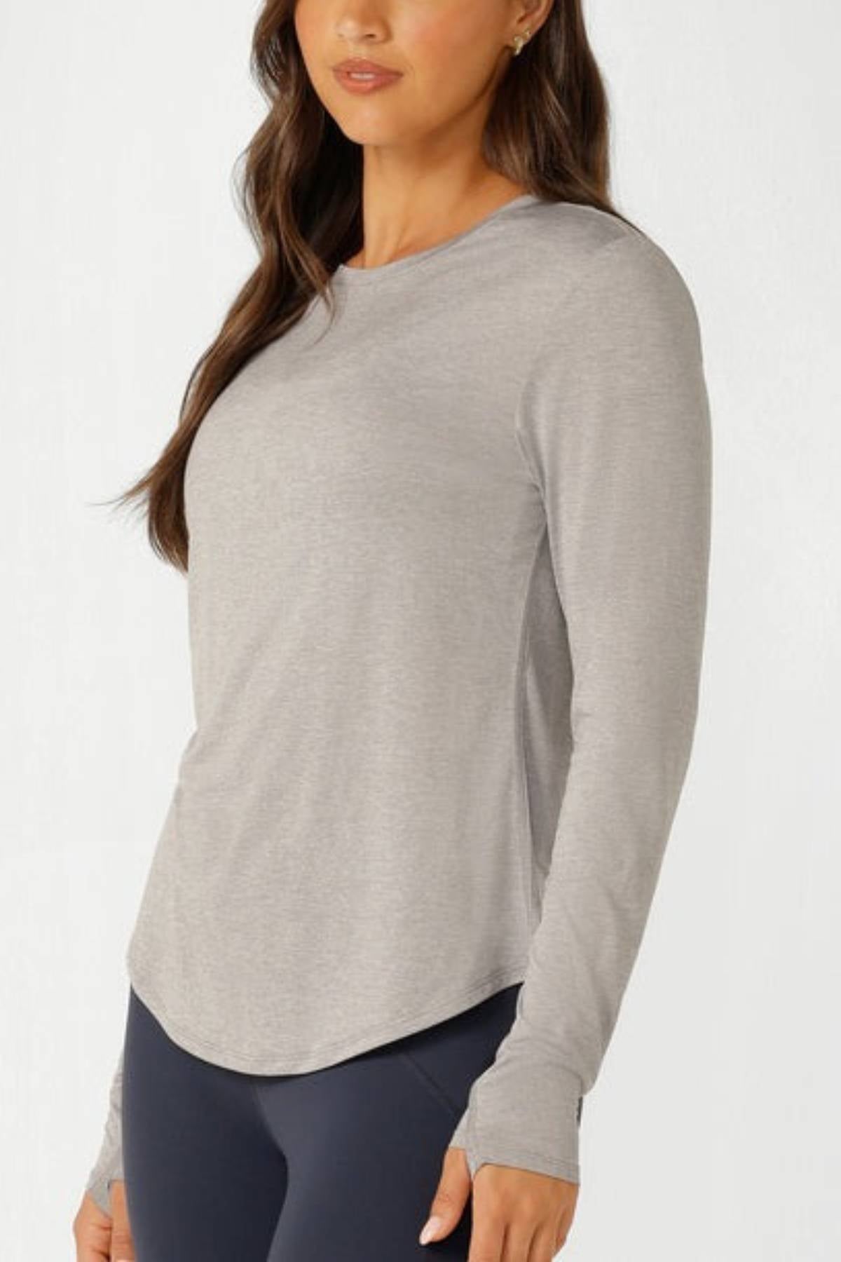 A woman wears the Lorna Jane Amy Dynamic Active Long Sleeve T-Shirt with dark leggings, standing against a white background and facing slightly to the side—ideal for lightweight layering.