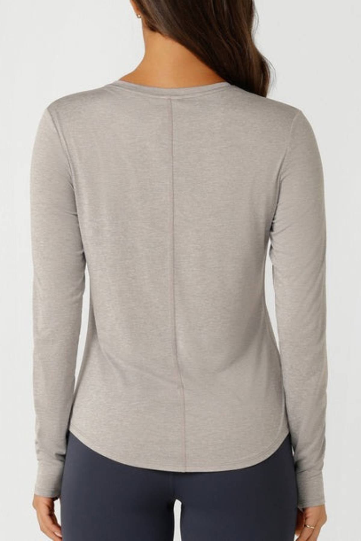 A woman with long brown hair, seen from the back, wears a Lorna Jane Amy Dynamic Active Long Sleeve T-Shirt and dark leggings against a plain light background—perfect for lightweight layering.