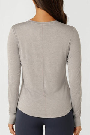 A woman with long brown hair, seen from the back, wears a Lorna Jane Amy Dynamic Active Long Sleeve T-Shirt and dark leggings against a plain light background—perfect for lightweight layering.