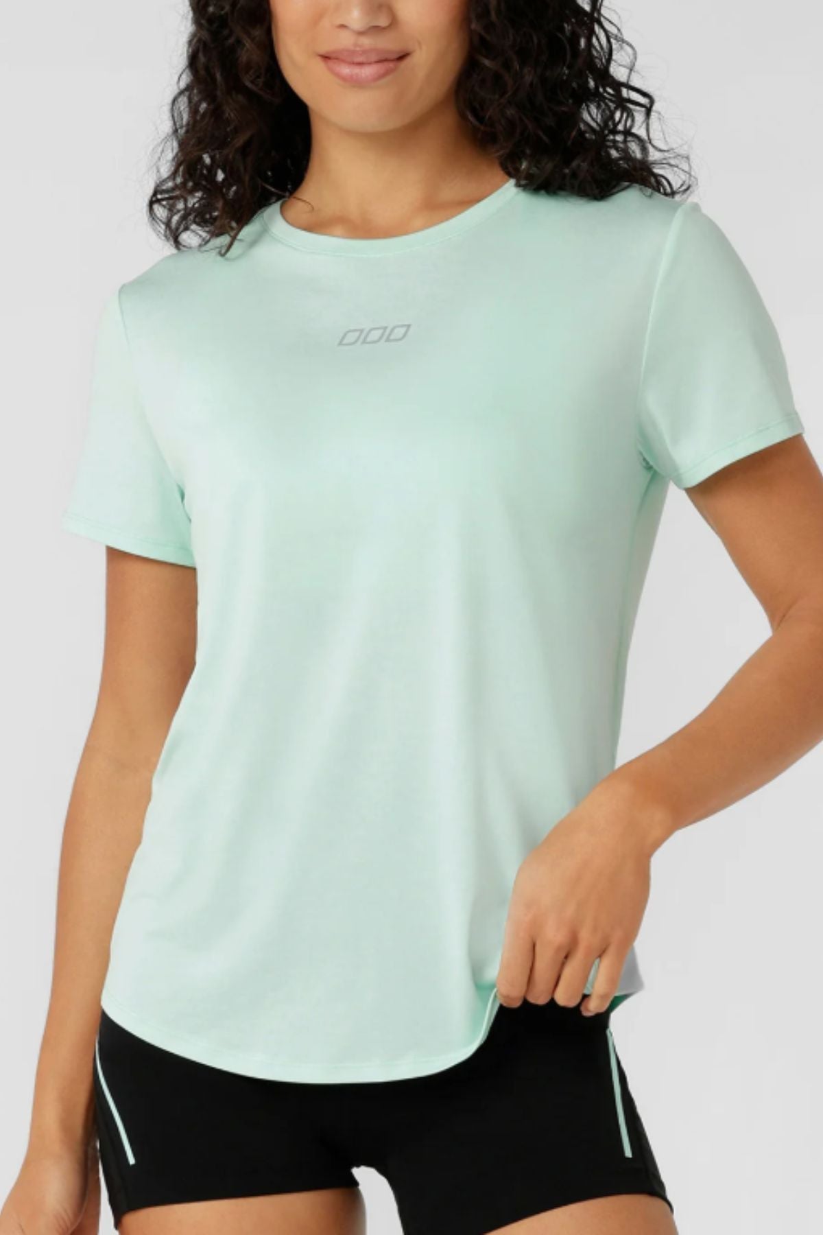 A woman with curly dark hair models the Lorna Jane Amy Dynamic Active T-Shirt in light green with black shorts, standing confidently against a plain backgroundβideal for those seeking a quick-drying women's workout top.