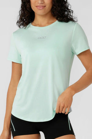 A woman with curly dark hair models the Lorna Jane Amy Dynamic Active T-Shirt in light green with black shorts, standing confidently against a plain backgroundβideal for those seeking a quick-drying women's workout top.