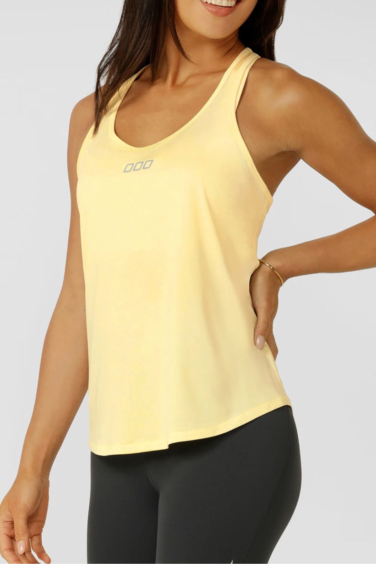 A woman stands smiling with one hand on her hip, wearing the Lorna Jane Amy Dynamic Active Tank in light yellow and black leggings against a plain background.