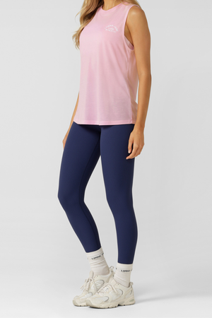 A woman wears a pink Lorna Jane Lotus Muscle Tank, navy blue leggings, white socks, and white sneakers, standing against a plain light background. Only her body from the shoulders down is visible.