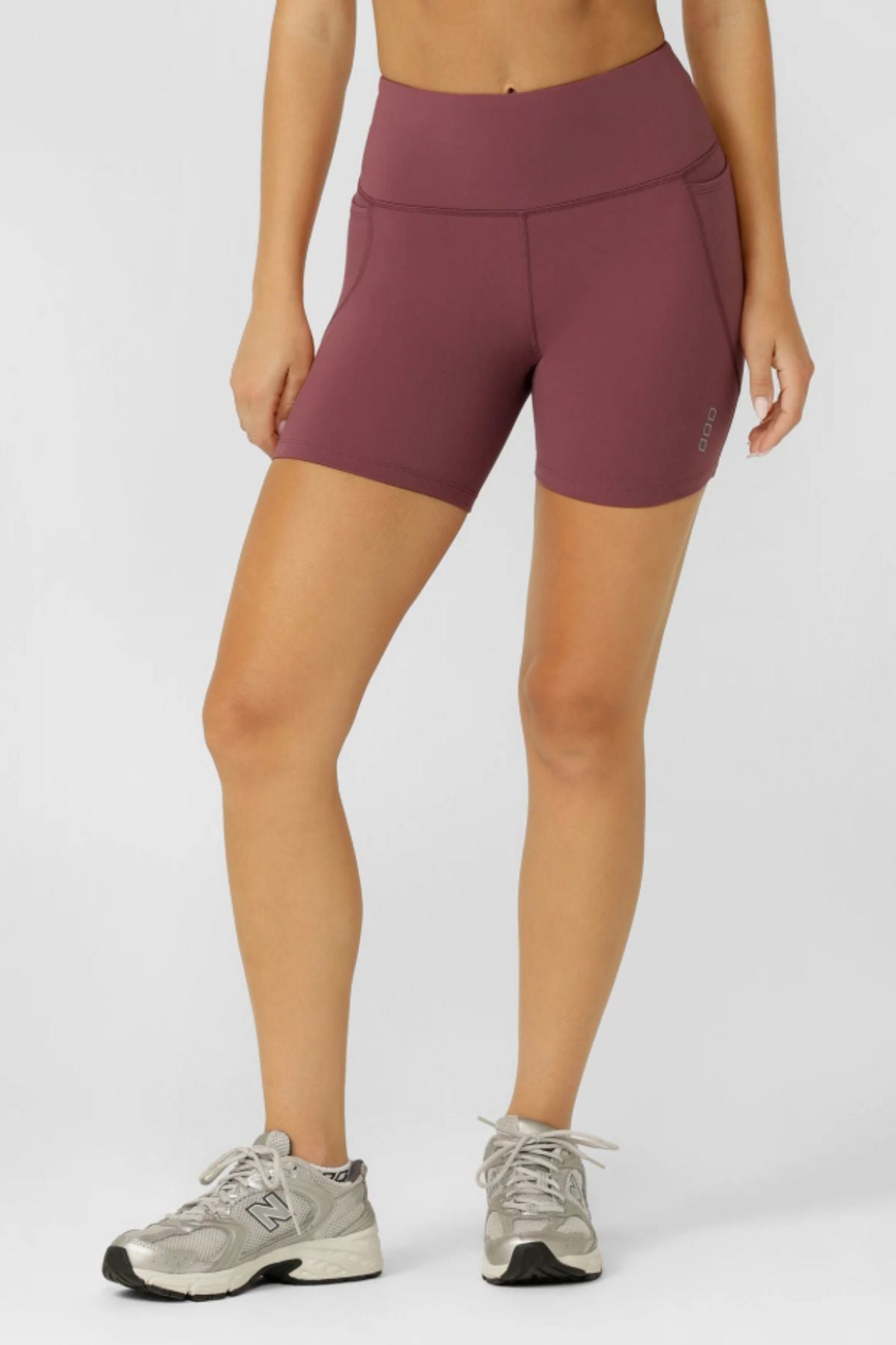 A person wears Lorna Jane Amy No Chafe Phone Pocket 14cm Bike Shorts in mauve and gray running shoes, standing against a plain light background with hands by their sides. Only the lower body is visible.