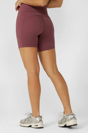 A person faces away, wearing mauve Lorna Jane Amy No Chafe Phone Pocket 14cm Bike Shorts with Active Core Stability and light gray sneakers. The plain background highlights the shorts’ sweat-wicking design.