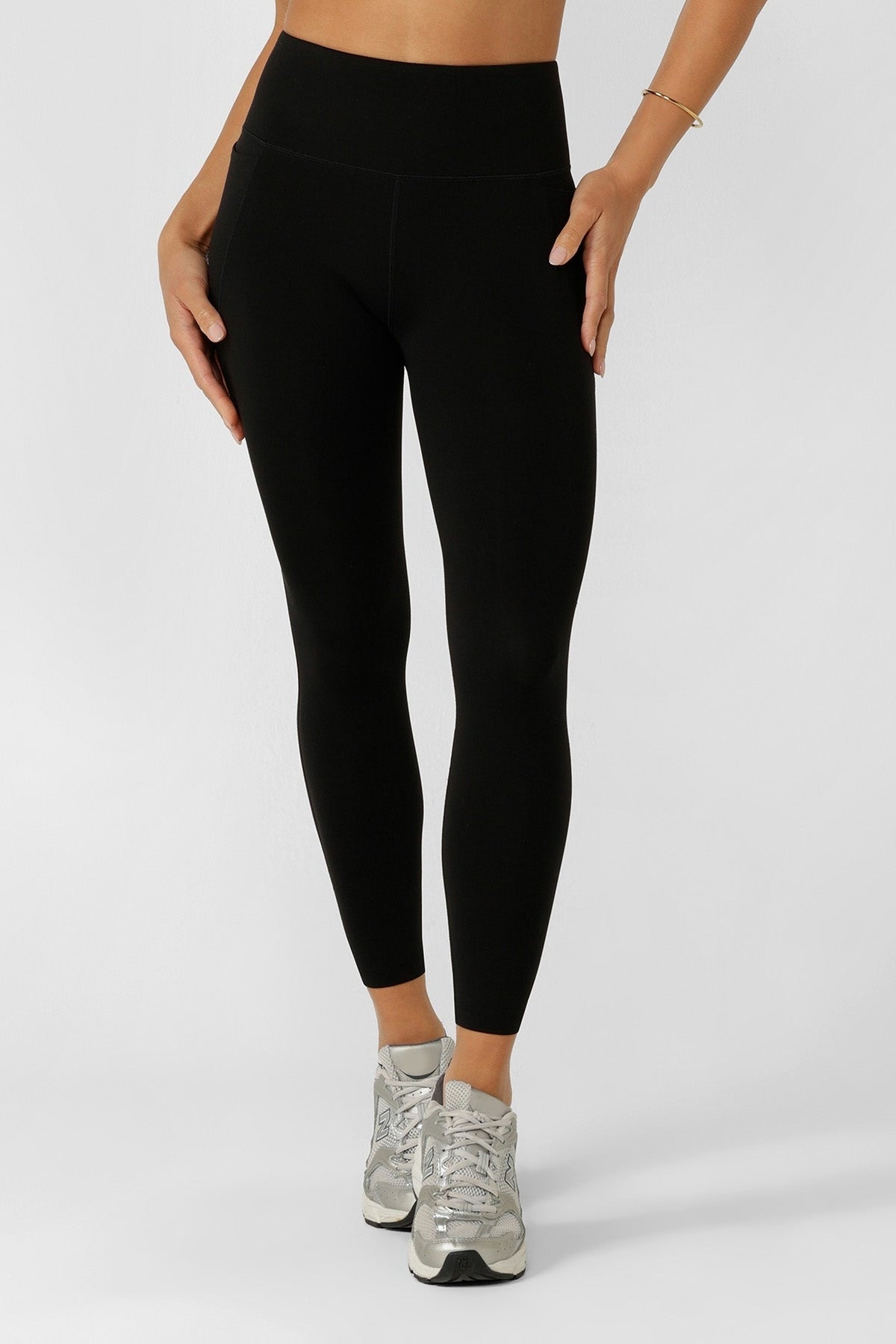 A person wears Lorna Jane Amy No Chafe Phone Pocket Ankle Biter Leggings and silver athletic shoes, standing against a light background with hands at their sides. Only the lower torso and legs are shown.