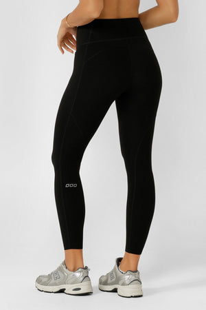 A person stands with their back to the camera in Lorna Jane Amy No Chafe Phone Pocket Ankle Biter Leggings, featuring a small white logo and sweat-wicking fabric, paired with silver athletic shoes against a plain light background.