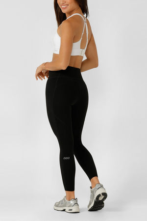 Woman smiling over her shoulder in a white sports bra, Lorna Jane Amy No Chafe Phone Pocket Ankle Biter Leggings with Active Core Stability, and silver sneakers, standing against a plain light gray background.