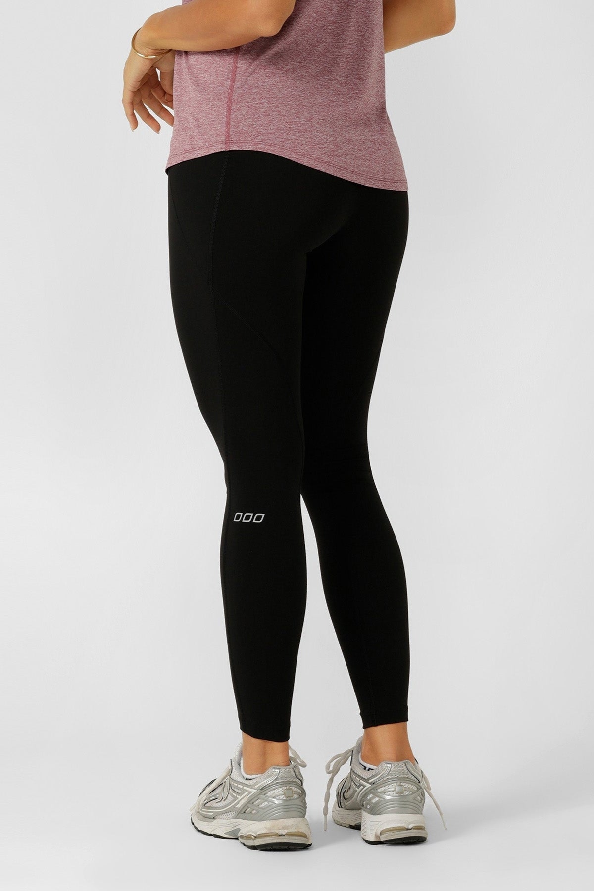 A person in a pink short-sleeve shirt and silver athletic shoes stands against a plain light background, wearing Lorna Jane Amy No Chafe Phone Pocket Full Length Leggings with 4-way stretch, body facing away and one hand on their hip.