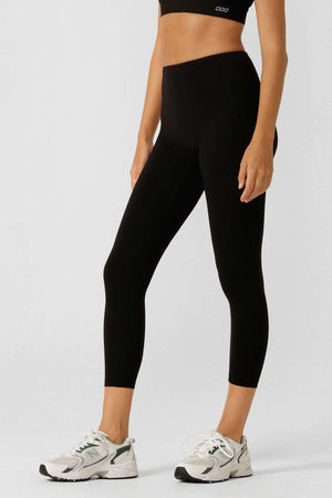 A person in a black crop top, white sneakers, and Lorna Jane Amy Phone Pocket 7/8 Tech Leggings with moisture-wicking fabric stands against a plain light background, arms relaxed at their sides.