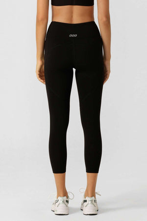 A person is shown from the back against a plain light background, wearing black Lorna Jane Amy Phone Pocket 7/8 Tech Leggings—made from moisture-wicking fabric—with a black sports bra and white sneakers.