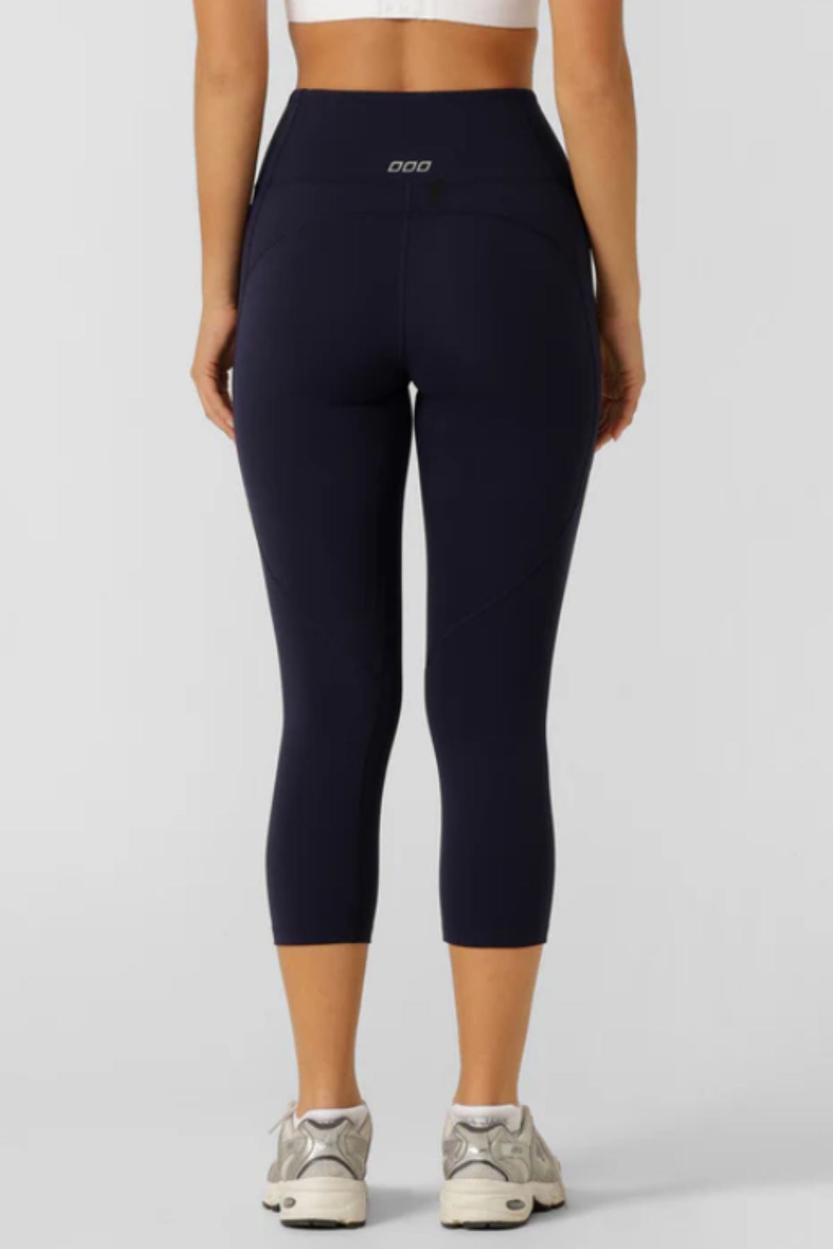 A woman faces away from the camera, wearing a white crop top and Lorna Jane Amy Phone Pocket 7/8 Tech Leggings—high-rise, moisture-wicking leggings—paired with white sneakers against a plain light background.