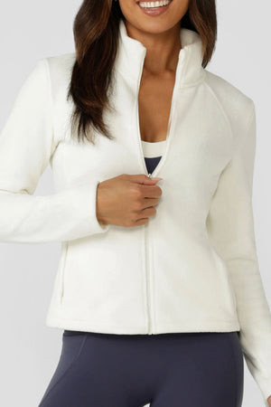 A woman smiles as she zips up the white Lorna Jane Amy Polar Fleece Zip Through Jacket, worn over a dark sports bra and navy leggings. The background is plain light gray.