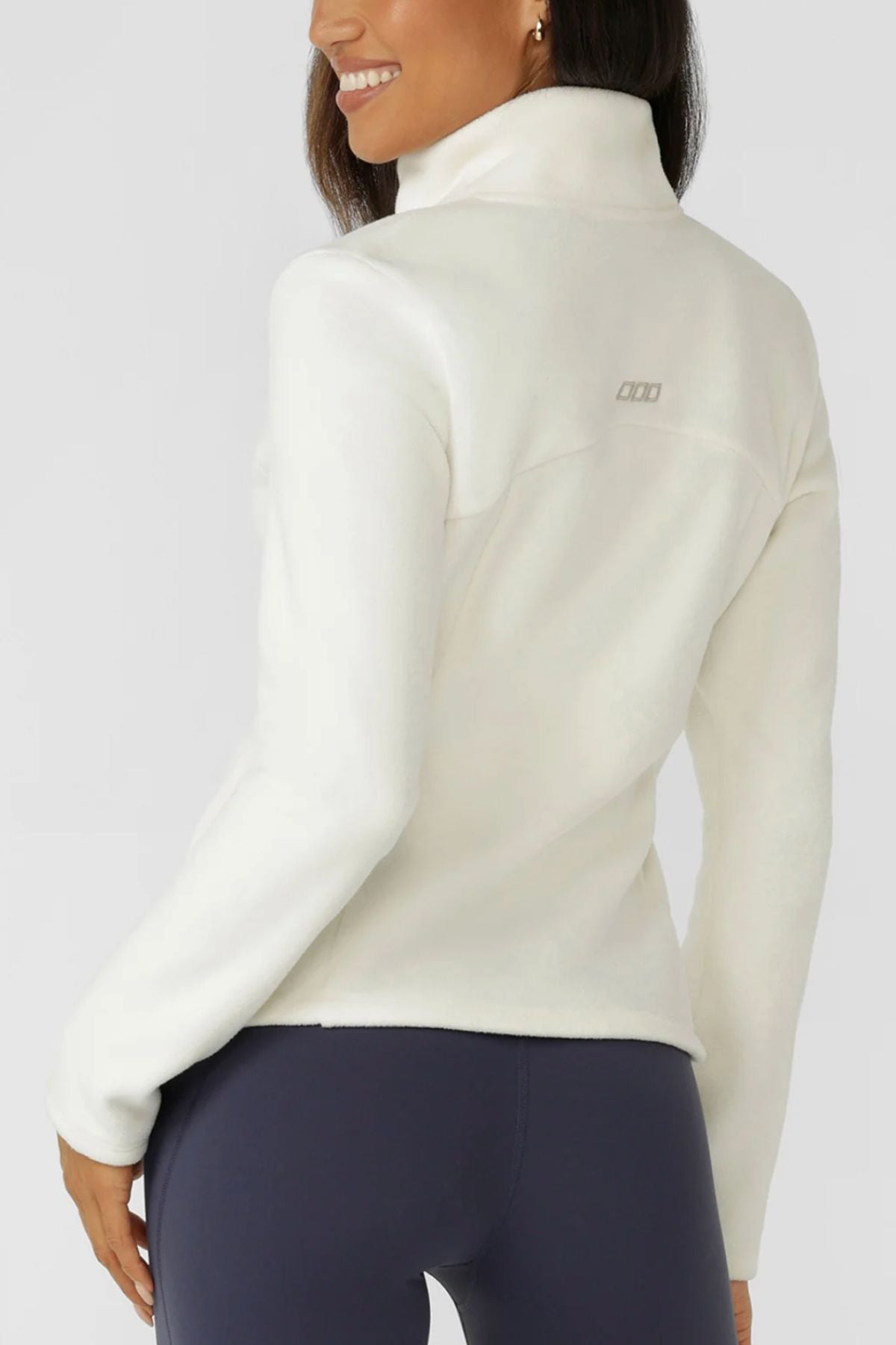 A woman smiles while wearing the Lorna Jane Amy Polar Fleece Zip Through Jacket and dark leggings, shown from the back against a light gray background.