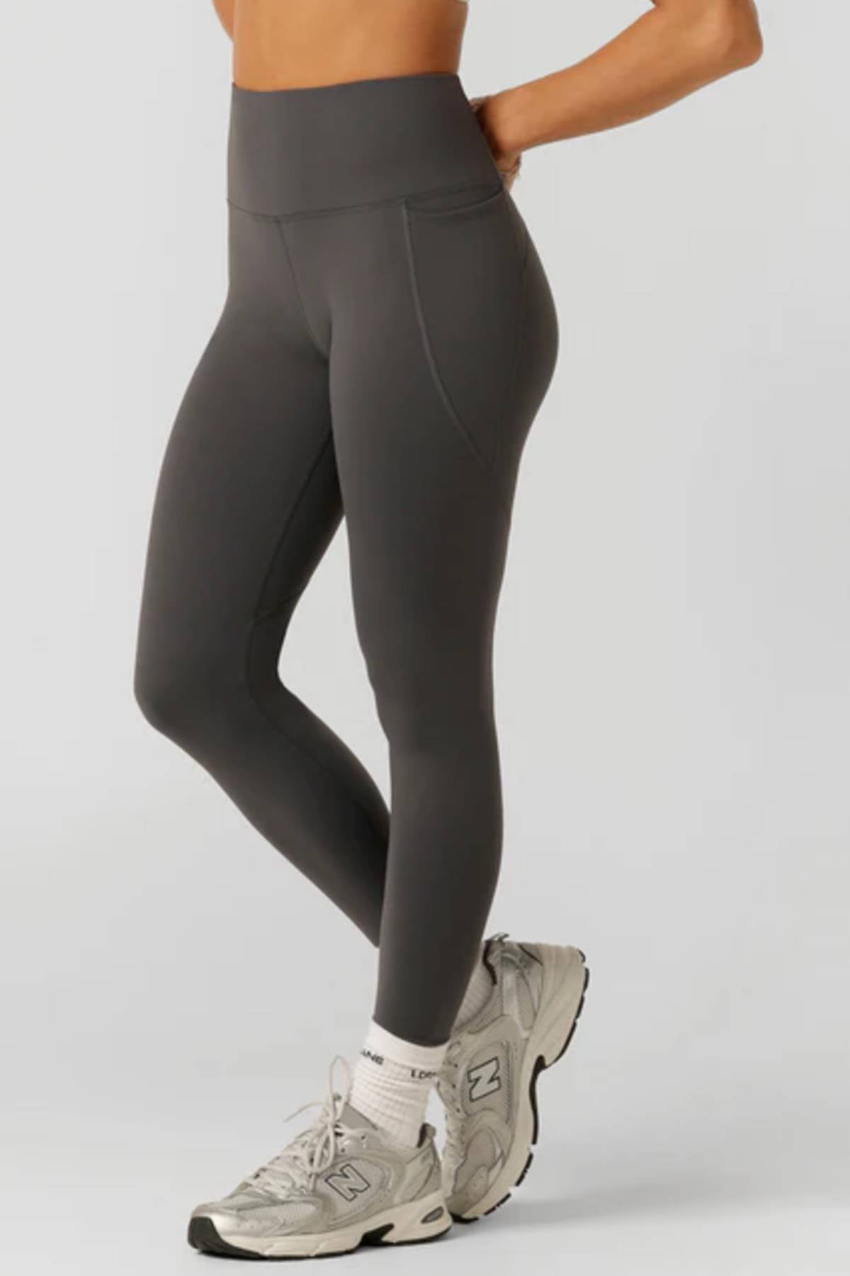 Wearing Lorna Jane Amy Thermal Phone Pocket Full Length Leggings in dark gray, paired with white crew socks and light gray New Balance sneakers. Only the lower body is visible against a plain light background.