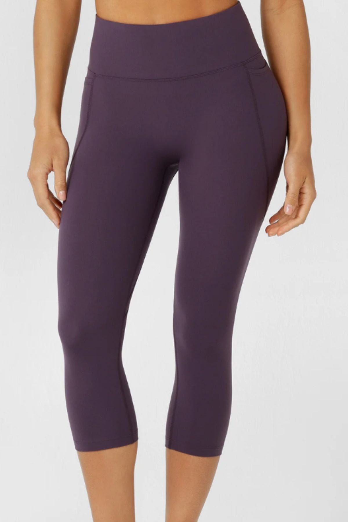 A person wears Lorna Jane Amy No Ride Phone Pocket 7/8 Leggings in dark purple, standing against a plain light background. Only their lower torso and legs are shown, emphasizing the high-rise fit and Active Core Stability design.