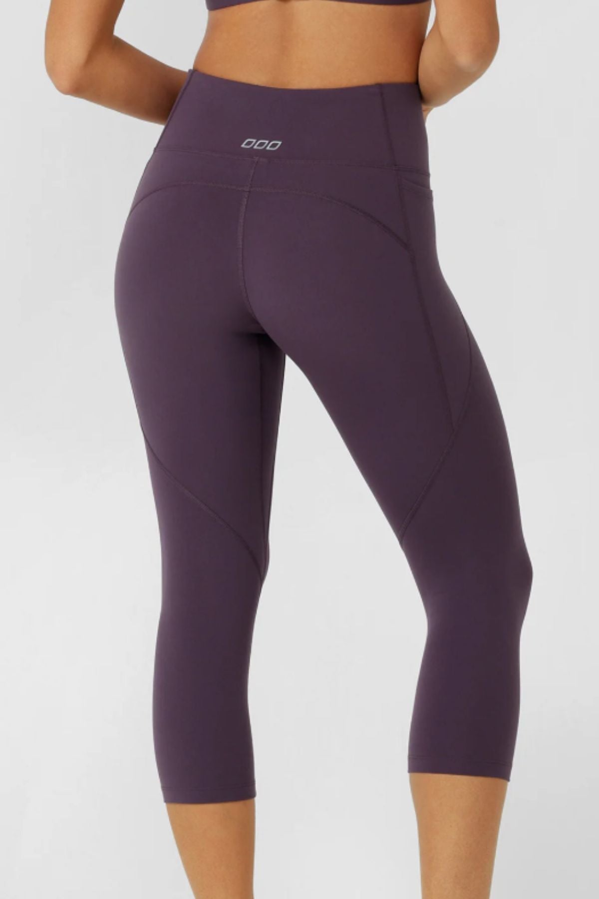 A woman is shown from the back wearing high-waisted, dark purple Lorna Jane Amy No Ride Phone Pocket 7/8 Leggings and a matching sports bra, standing against a plain light gray background.