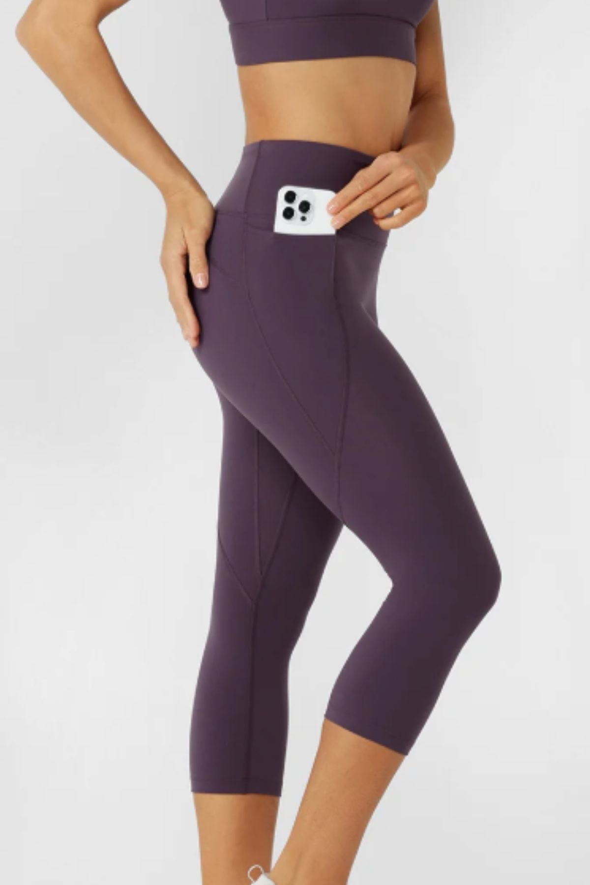 Wearing Lorna Jane Amy No Ride Phone Pocket 7/8 Leggings, a person stands against a plain background, matching a purple sports bra and tucking a phone into the leggings' waistband pocket.