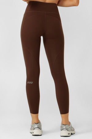A person is shown from the back wearing Lorna Jane Amy No Chafe Phone Pocket Ankle Biter Leggings in brown with sweat-wicking fabric and gray sneakers, standing against a plain light-colored background.