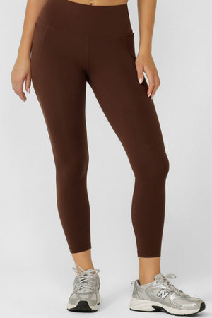 A person wears Lorna Jane Amy No Chafe Phone Pocket Ankle Biter Leggings and gray New Balance sneakers, standing against a plain white background with one hand by their side and the other on their thigh.