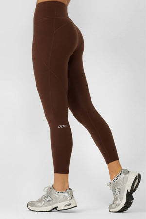 Someone wearing Lorna Jane Amy No Chafe Phone Pocket Ankle Biter Leggings in high-waisted brown, made from sweat-wicking fabric, and silver New Balance sneakers stands sideways on a neutral background with one leg slightly forward.