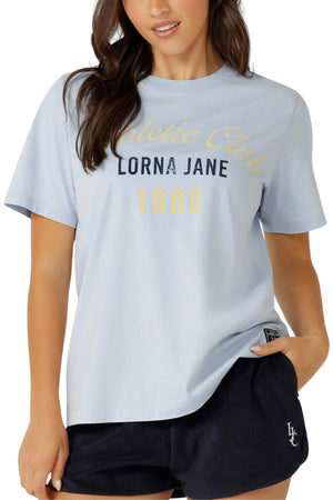 A woman stands with one hand in her pocket, wearing a relaxed-fit Lorna Jane Athletic Club Relaxed T-Shirt in light blue and dark blue shorts.