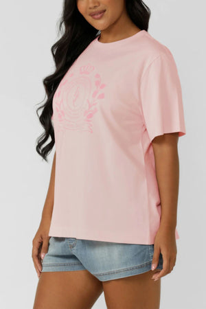 A woman in the Lorna Jane Chalet Relaxed T-Shirt—premium cotton, light pink, with a subtle front crest—paired with light blue denim shorts, stands against a plain background, embodying an active lifestyle.