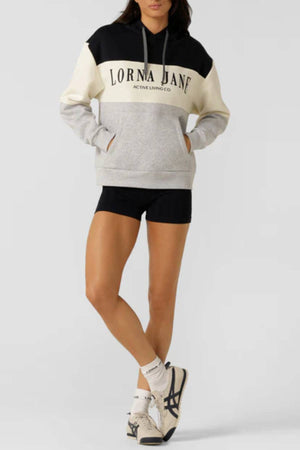 Person wearing a Lorna Jane Championship Oversized Fleece Hoodie with color blocking, black shorts, white socks, and white sneakers stands with one leg crossed over the other against a plain light background.