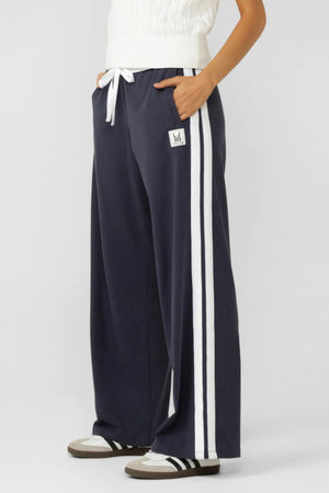 A person wears Lorna Jane Dance Pants in navy blue with a white adjustable waistband and side stripes for a retro sport vibe, paired with a white knit top and black-and-white sneakers.
