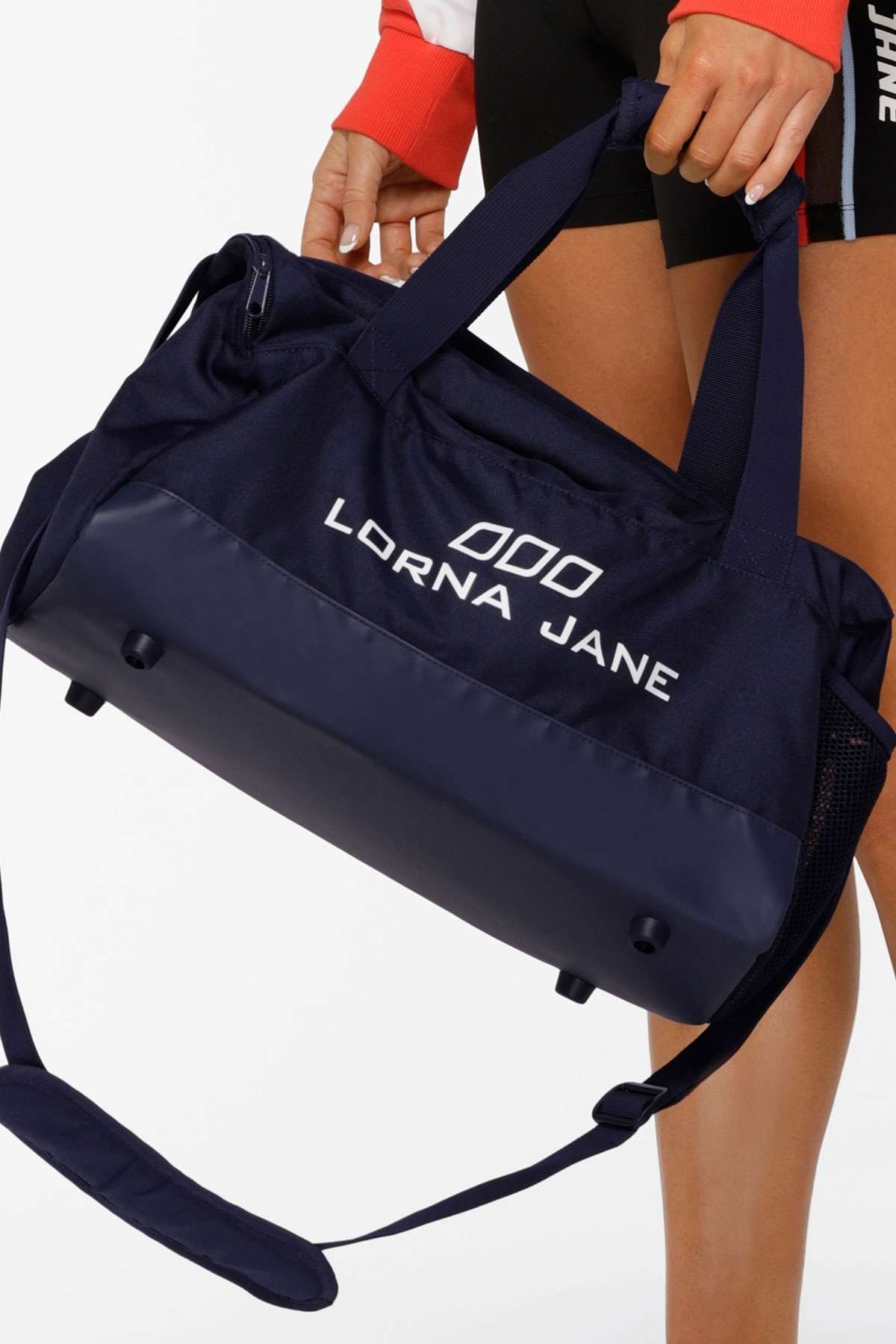 A person holds the Lorna Jane Essential Gym Bag in navy, featuring a multi-pocket design, dual handles, and an adjustable shoulder strap, while wearing black shorts and a red and white jacket.