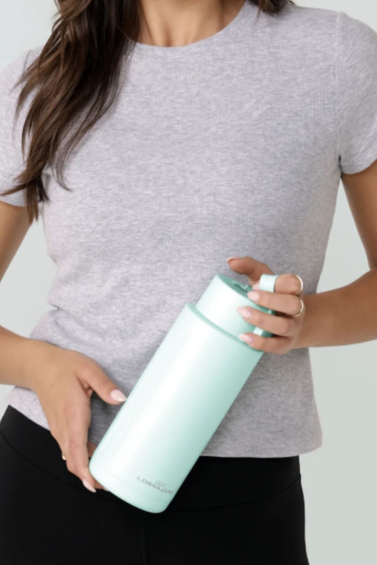 A woman in a light grey t-shirt and black pants holds a large, light blue Lorna Jane Essential Insulated Water Bottle (1L) with a handle near the cap.