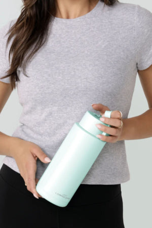 A woman in a light grey t-shirt and black pants holds a large, light blue Lorna Jane Essential Insulated Water Bottle (1L) with a handle near the cap.