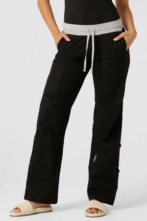 A person wears Lorna Jane Flashdance Pant in black with a gray waistband and adjustable elastic waist for comfort, paired with white slide sandals, standing hands-in-pockets against a plain background.