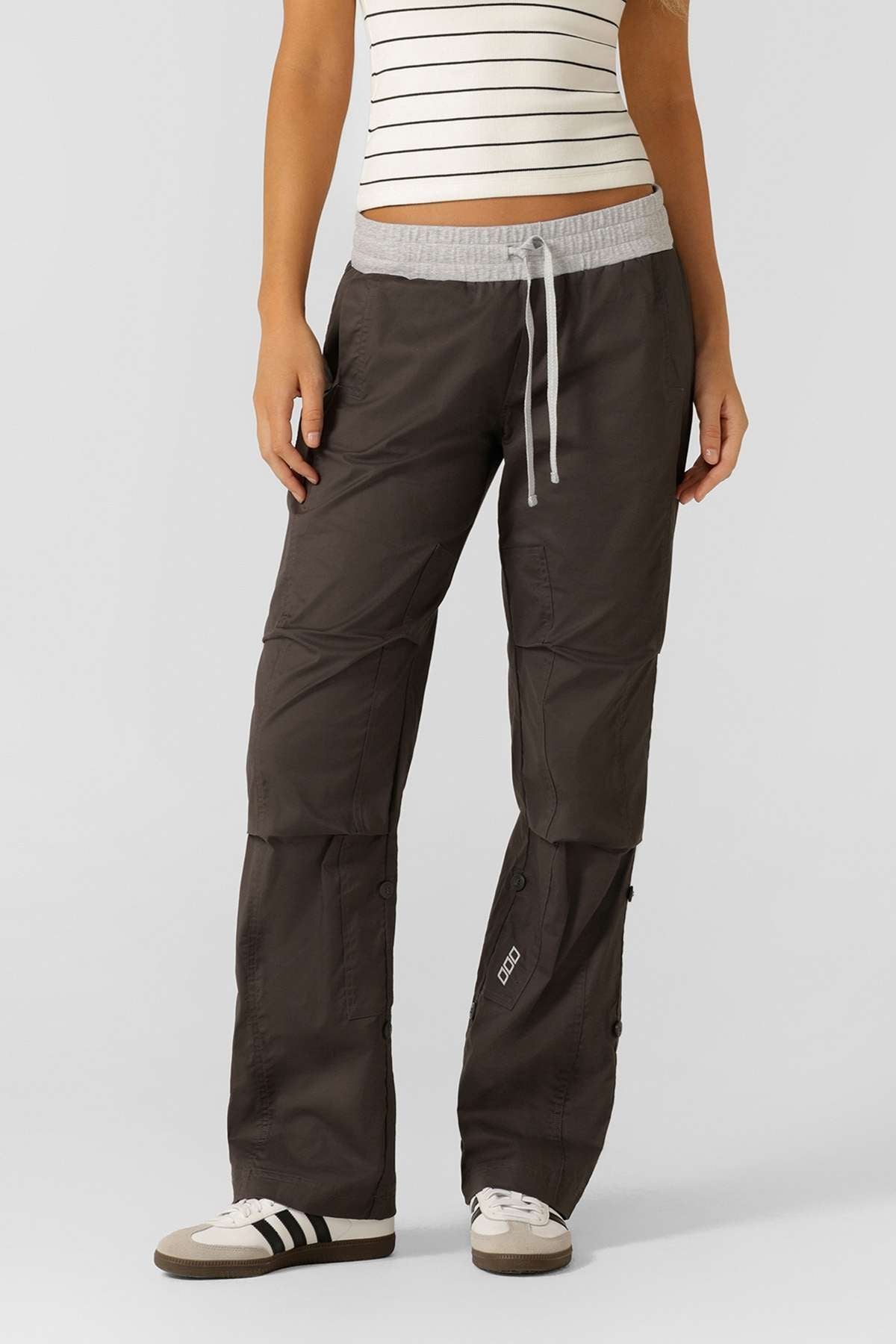 A person wears a white and black striped tank top, gray Lorna Jane Flashdance Pant cargo pants with reinforced seams, and sneakers, standing against a plain light background.