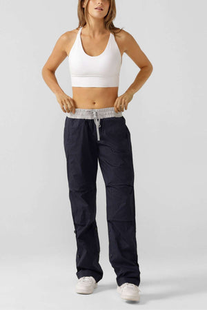 A woman stands against a plain background wearing a white sports bra, the Lorna Jane Flashdance Pant in loose dark navy with reinforced seams, and white sneakers. She has long hair and is adjusting the waistband of her pants.