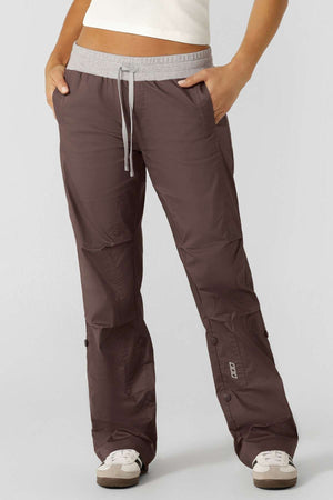 A person wearing Lorna Jane Flashdance Pant in brown, featuring an adjustable elastic waist, paired with white sneakers and standing with one hand in their pocket against a plain background.