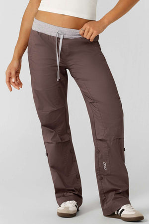 A person wears brown Lorna Jane Flashdance Pants with an adjustable elastic waist and white athletic shoes, cropped at the torso and ankles, standing against a plain background.