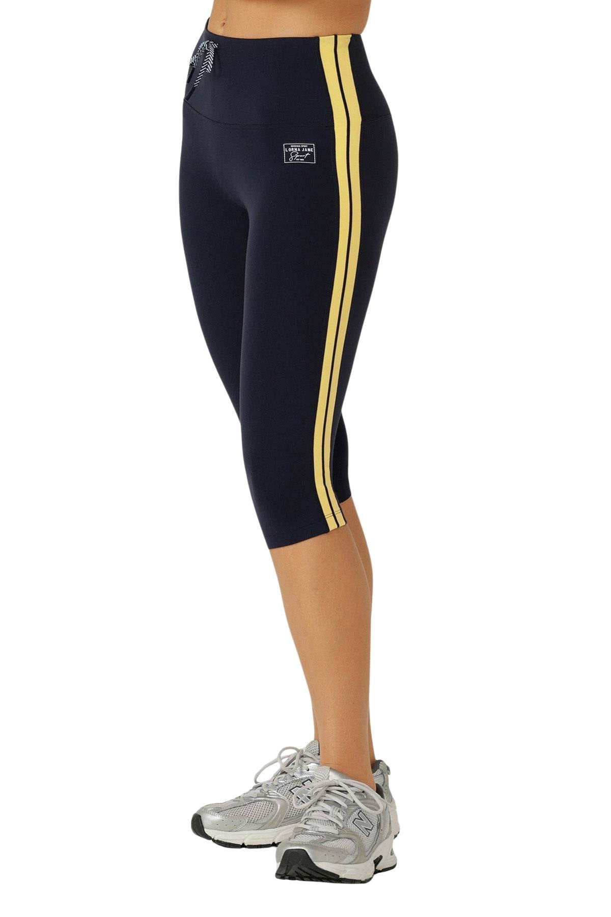 Wearing Lorna Jane Game Day 3/4 Capri Leggings in navy with yellow side stripes and gray athletic sneakers, a person stands on a white background, showcasing a retro sport style.