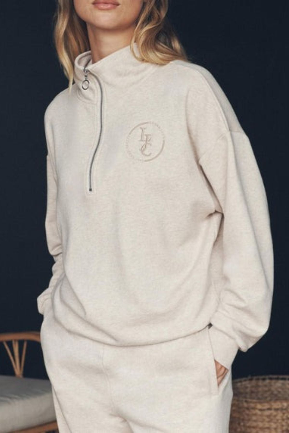 A woman stands against a dark background wearing the Lorna Jane LJC Heritage Quarter Zip Sweat in light beige with a small embroidered chest logo, matching sweatpants, and her hand in her pocket.