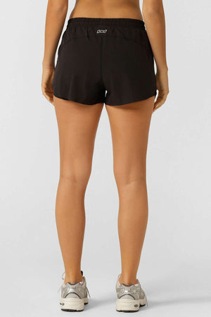 A person stands facing away in black Lorna Jane Lightspeed Phone Pocket Run Shorts with an elastic waistband, small white back logo, and sweat-wicking fabric, paired with a beige top and gray sneakers against a plain light background.