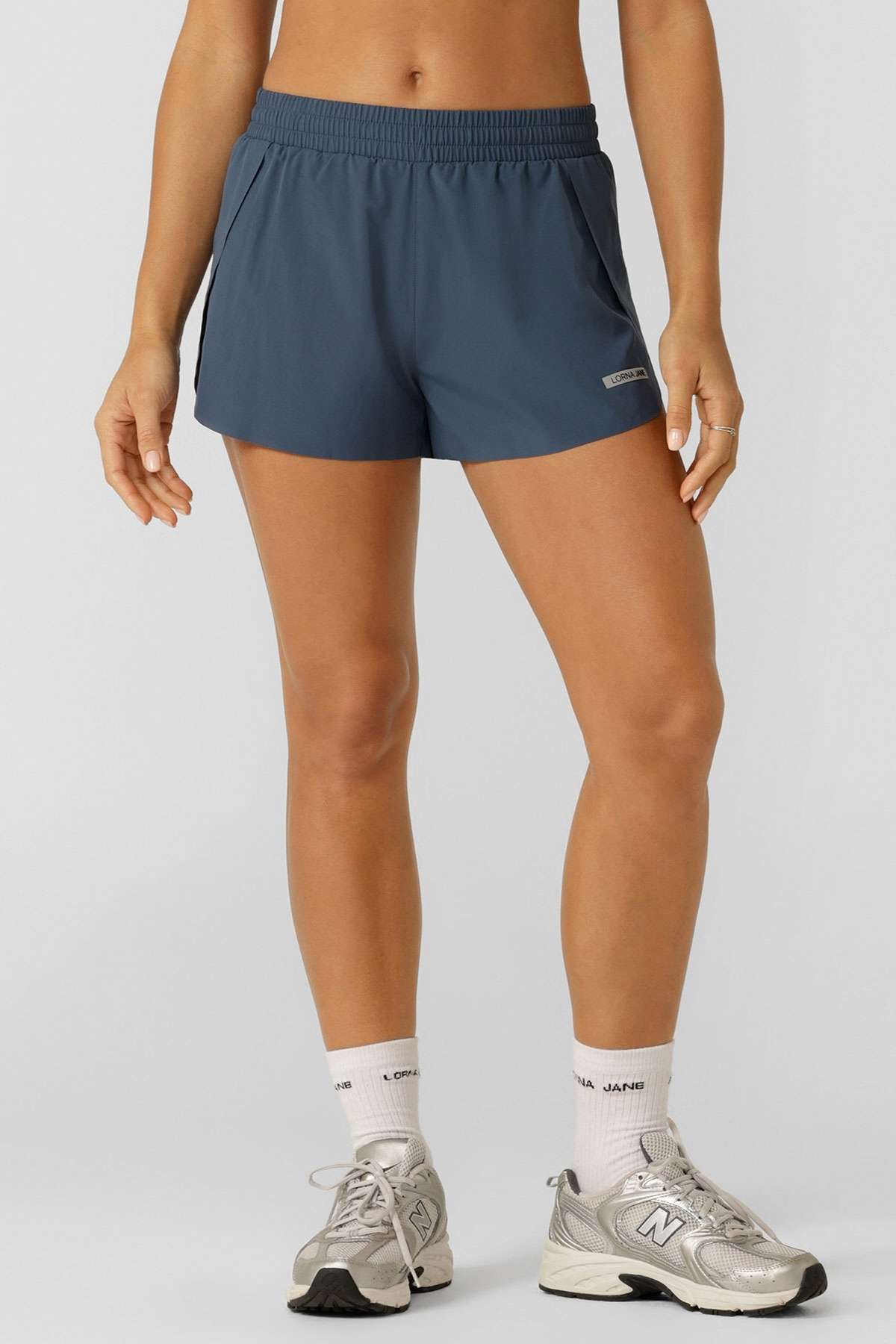 Someone stands against a light background, showing only their lower body in blue Lorna Jane Lightspeed Phone Pocket Run Shorts with an anti-chafe lining, white socks, and silver New Balance sneakers. Their hands rest at their sides.